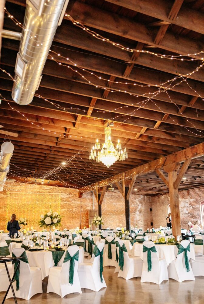 Wedding reception at Historic John B. Busch Brewery with string lights, exposed wood beams, and tall white floral centerpieces