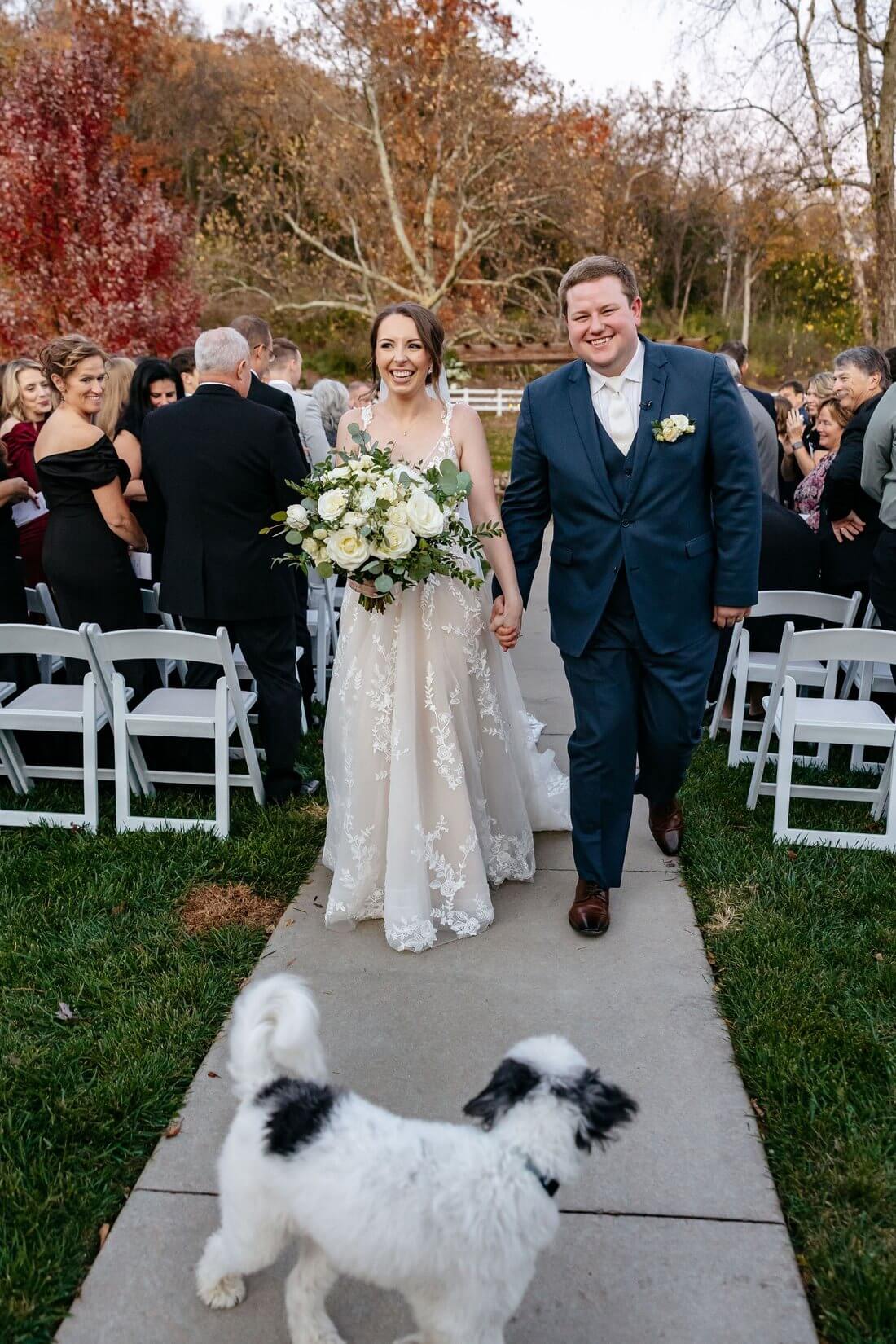 Newlyweds carrying a white rose bouquet while their puppy waits at the end of the aisle