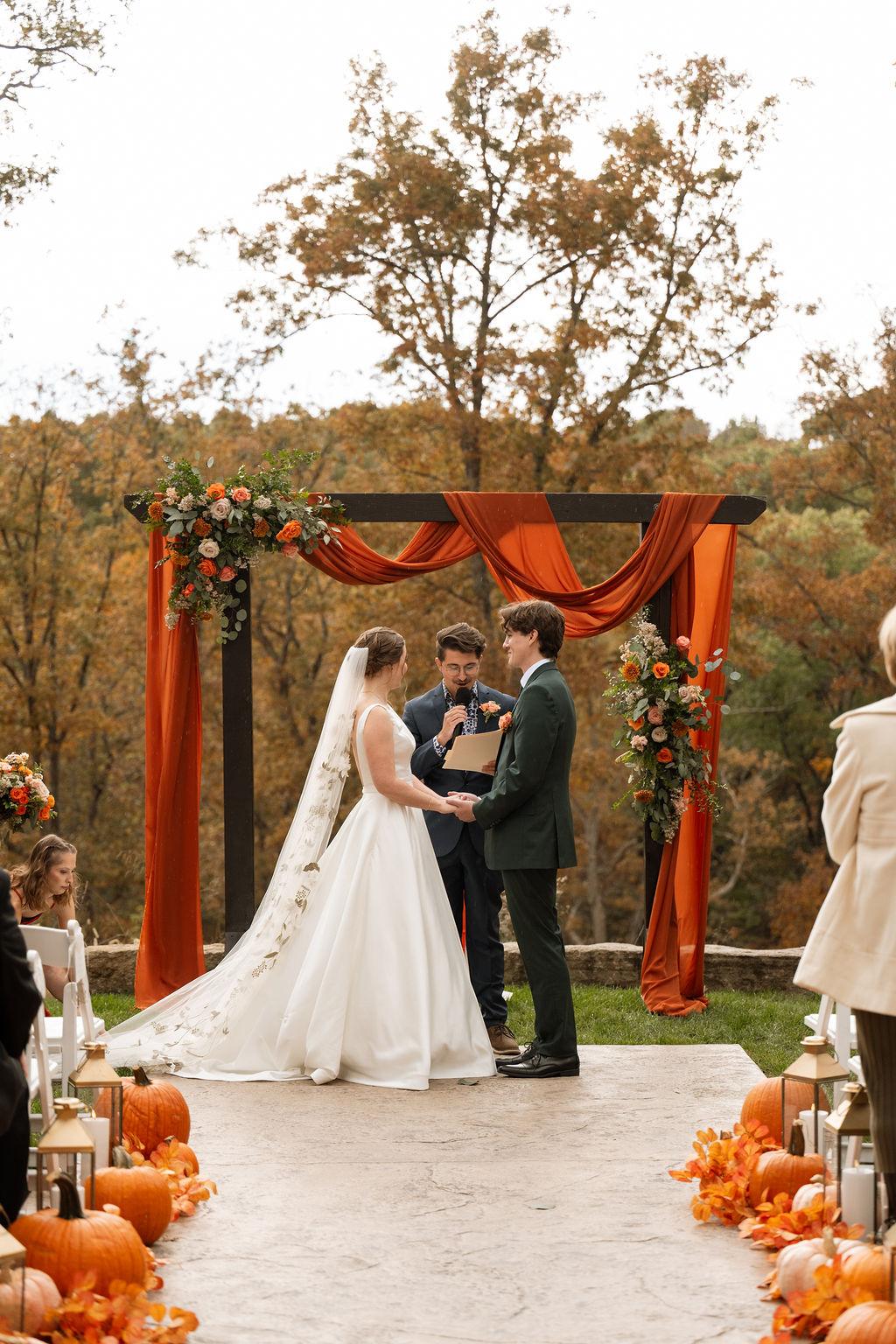 Bride and groom beneath a rust-draped floral arbor during a fall wedding ceremony at Silver Oaks Chateau